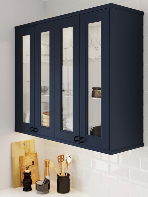 AXSTAD blue shaker wall cabinets with tall glass doors, in a bright kitchen with white worktop and white tiles.