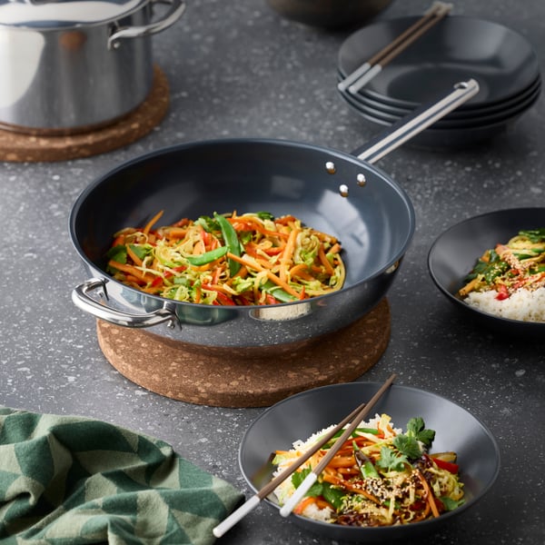 A kitchen featuring a stainless steel IKEA 365+ wok, filled with colourful sautéed vegetables, on a cork mat.