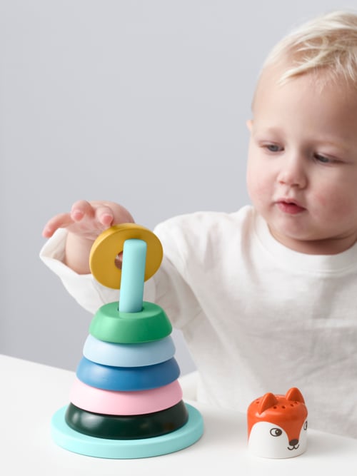 Multicoloured stacking rings are stacked on a blue stick, and a child is holding a yellow stacking ring.