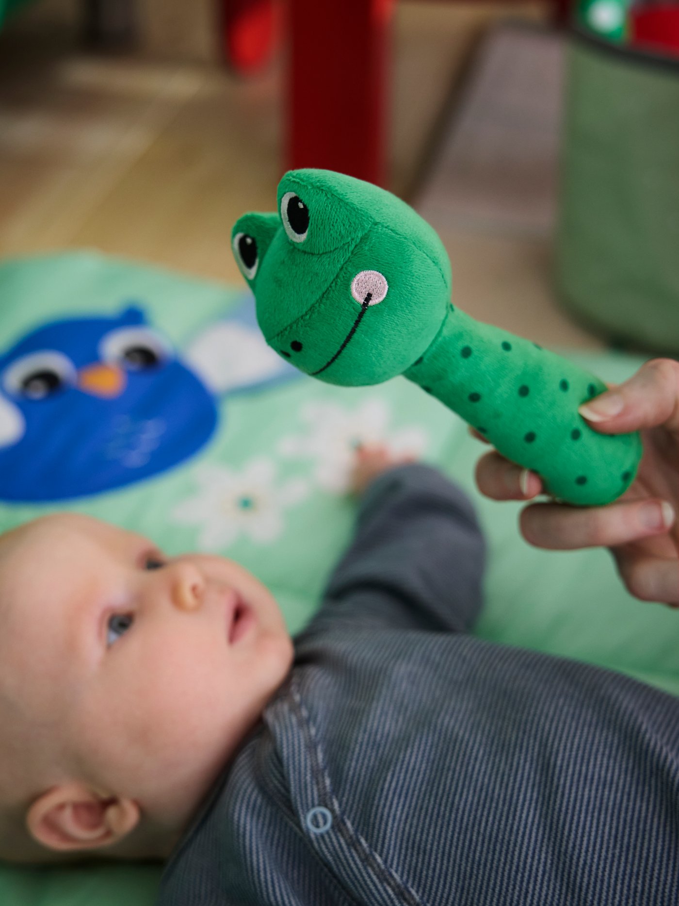 Close-up of baby laying on SANDBI play mat, with a grownup holding SANDBI bright green frog rattle close to the camera.