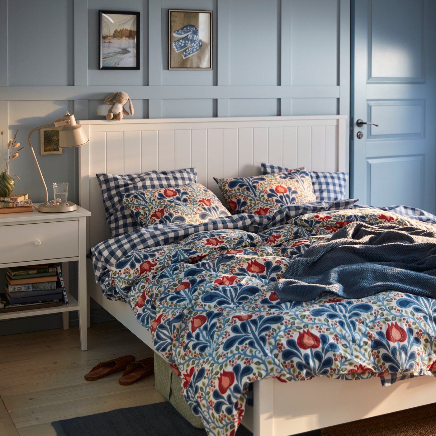 Un dormitorio pintado de azul con un juego de funda nórdica SÄTERVIDE con estampado floral en una cama blanca, junto a una mesilla de noche blanca en la que hay una lámpara.