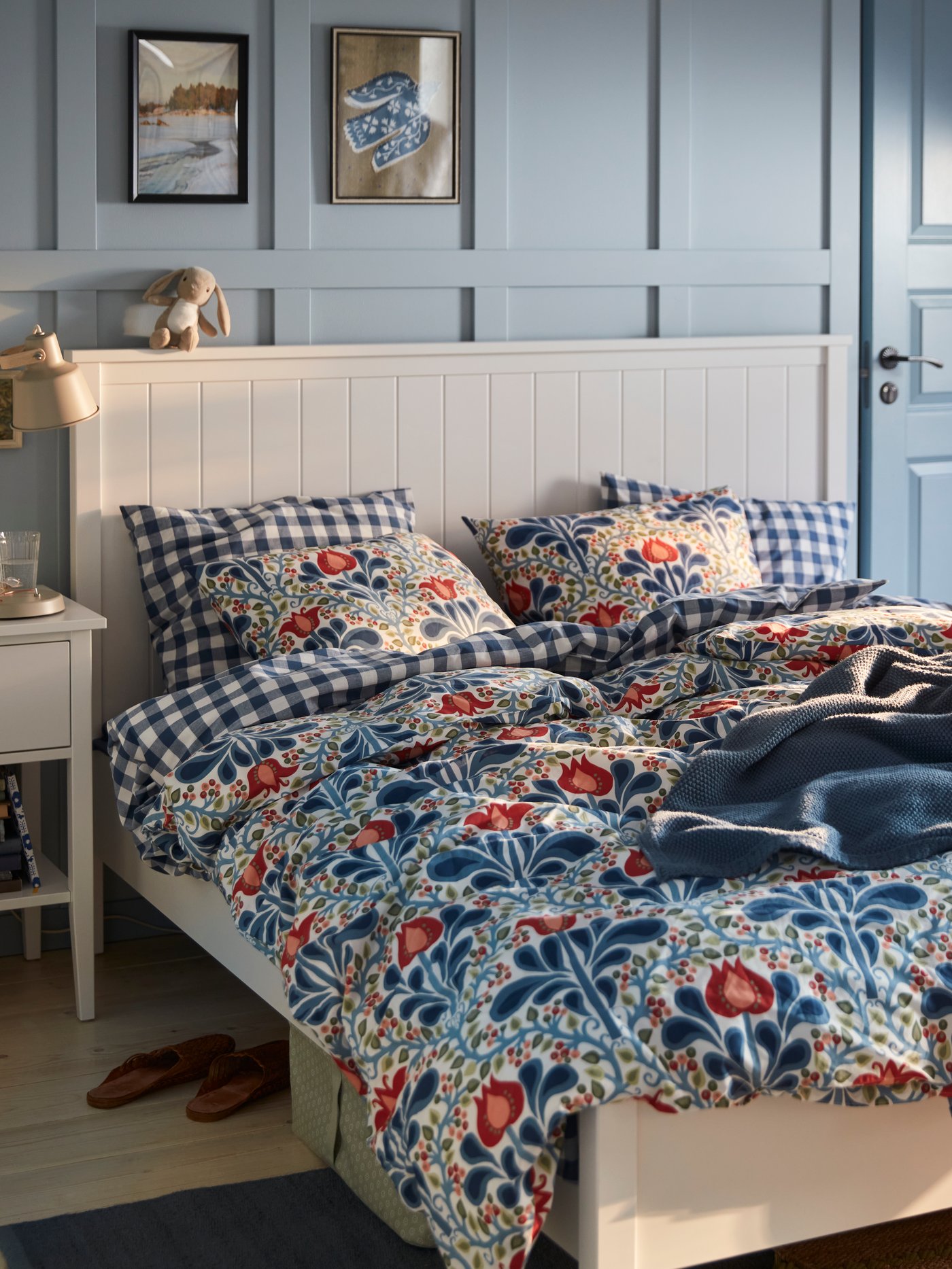 A blue painted bedroom with a floral SÄTERVIDE duvet cover set on a white bed, next to a white bedside table with a lamp.