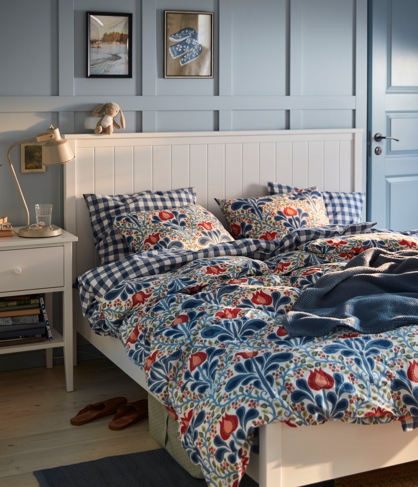 A blue painted bedroom with a floral SÄTERVIDE duvet cover set on a white bed, next to a white bedside table with a lamp.