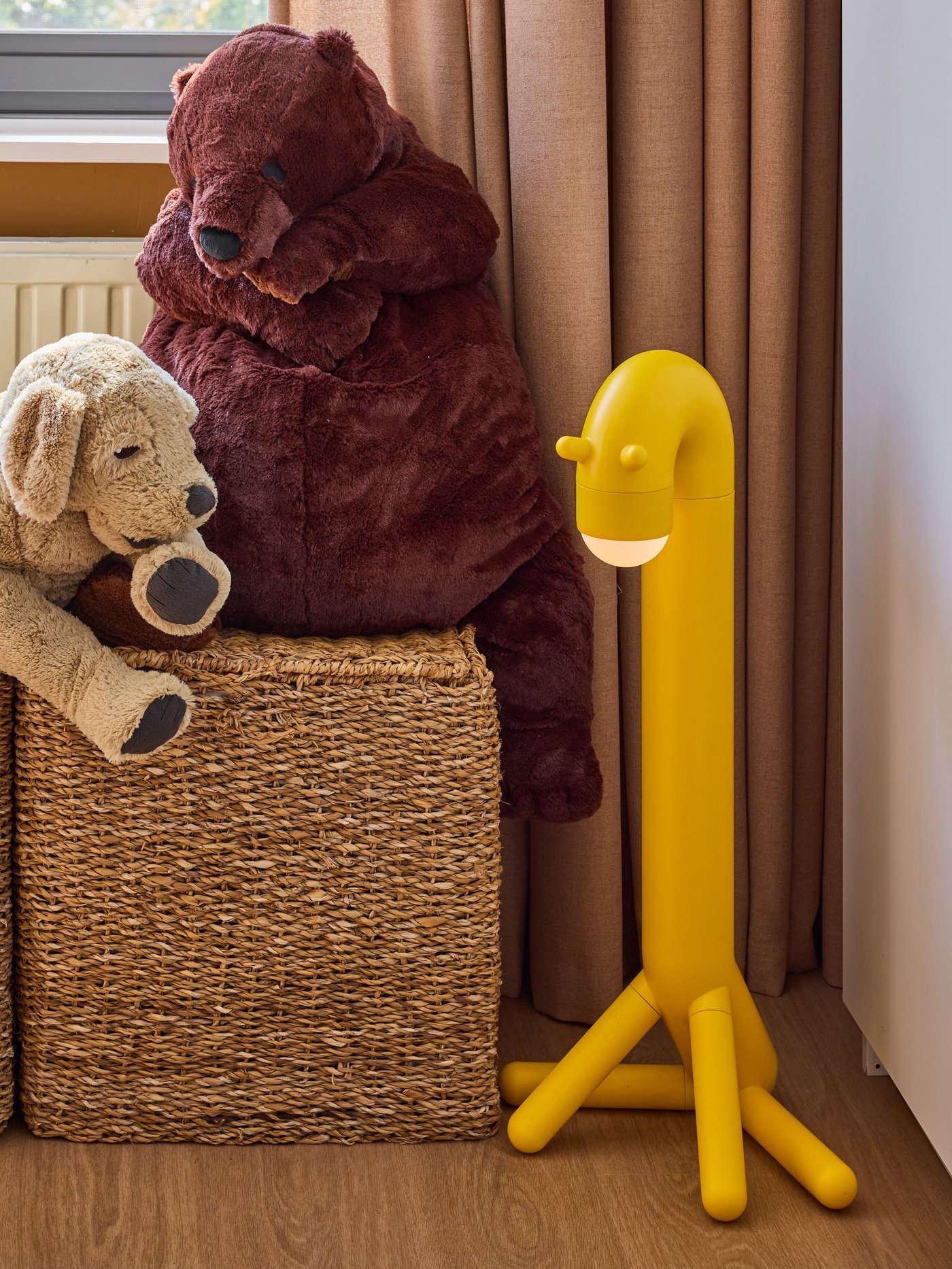 Gevlochten opbergmand met knuffels naast een gele kinderlamp in giraffevorm, tegen gordijnen in een warme kinderkamer.