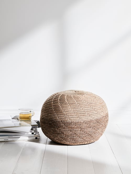 A SANDARED pouffe with a knitted beige cover is on a wooden floor by a white wall and stacked books with a cup of tea on top.