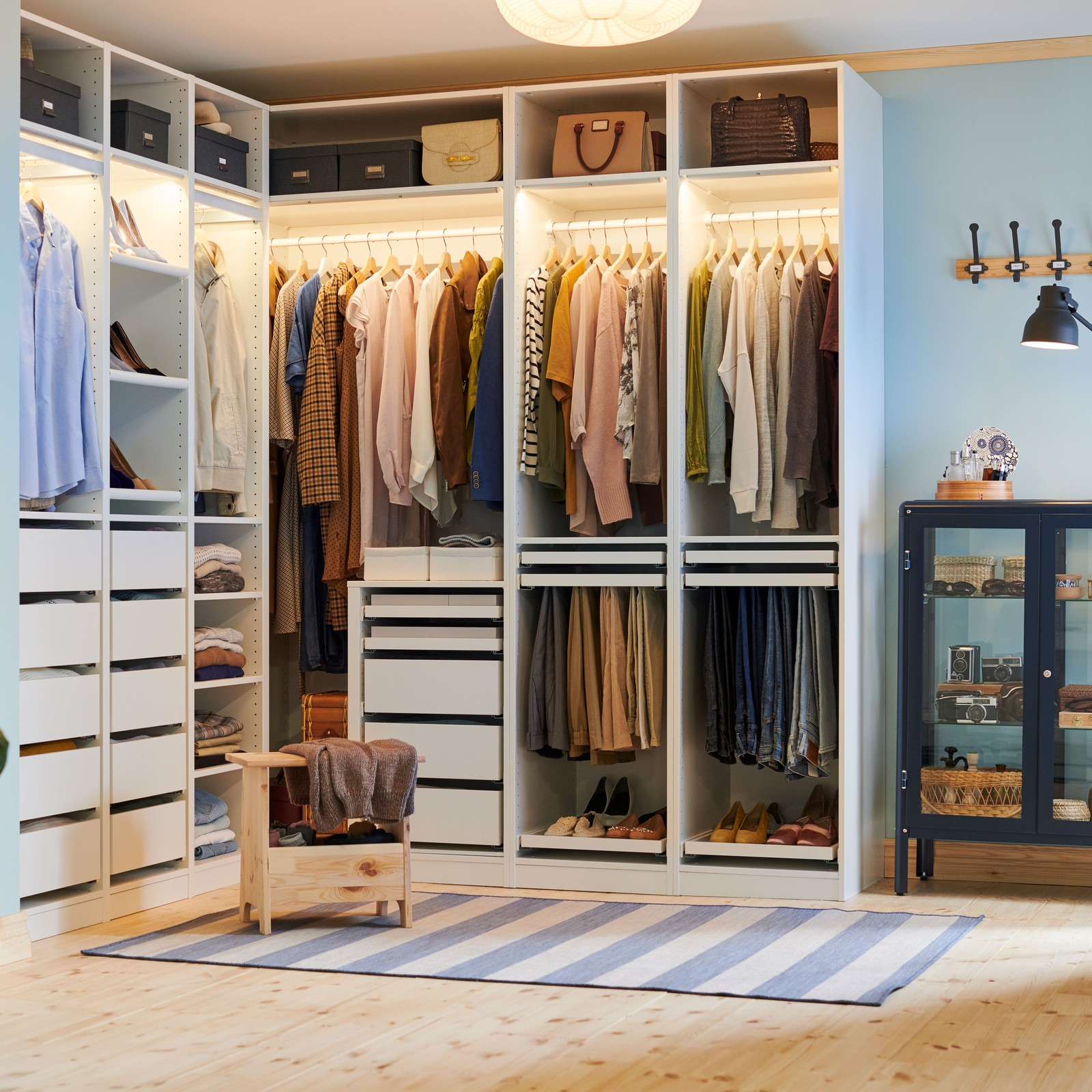 שרפרף עם אחסון PERJOHAN מעץ אורן ניצב מול ארון בגדים פינתי PAX פתוח וגדול שבו בגדים, נעליים, קופסאות ותיקים.