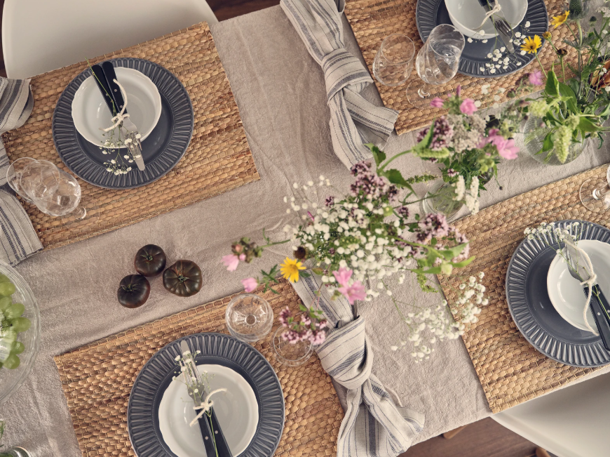 Obenansicht eines gedeckten Tisches mit vier Plätzen, geflochtenen Platzmatten, Geschirr in Blau‑Weiss und Wildblumen als Dekoration.