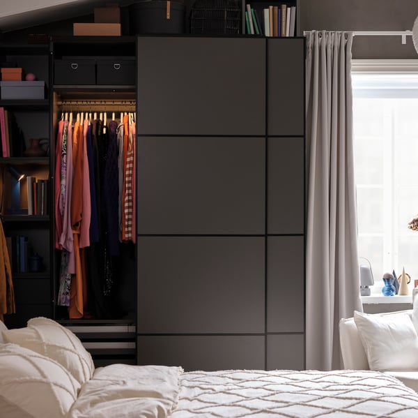 Behind a bed is a dark grey PAX/MEHAMN wardrobe with the left sliding door slightly open, showing clothes hanging on a rail.