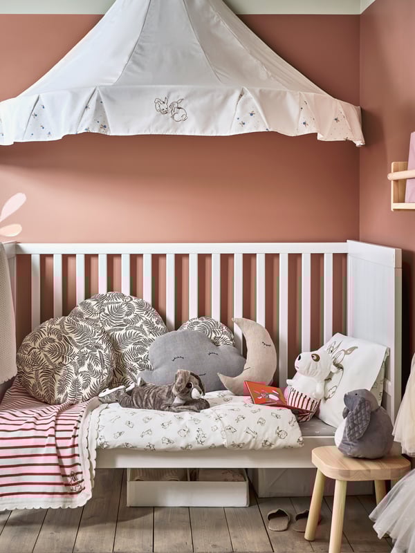 A children’s room where a SUNDVIK crib with cozy cushions, RÖDHAKE bed linen and soft toys sits under a white bed canopy.