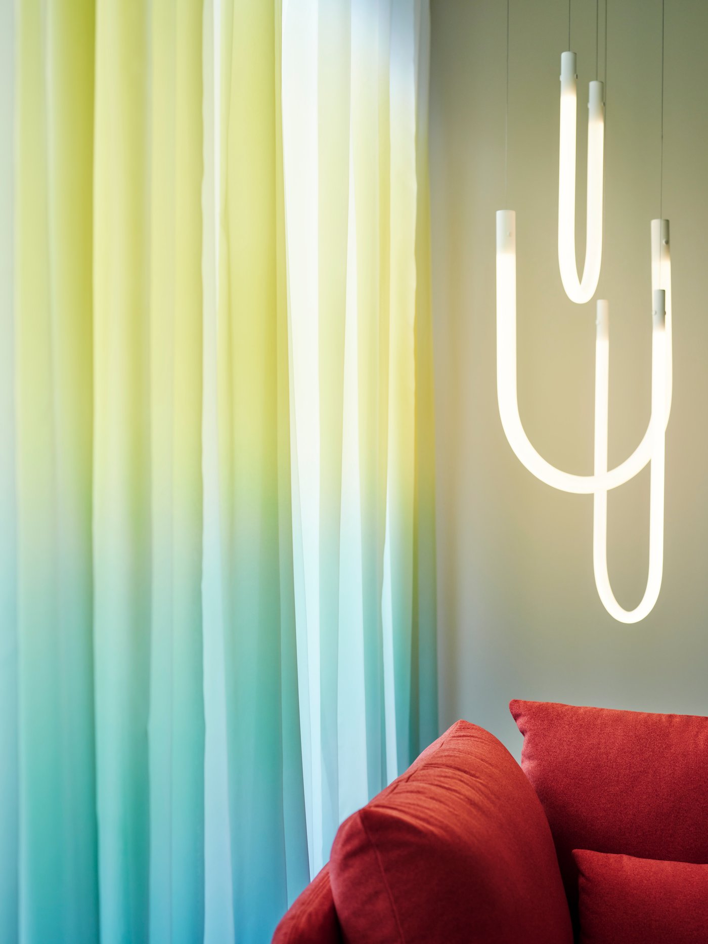 Two GLASÖRT sheer curtains hang in a window near a VARMBLIXT LED pendant lamp hanging from the ceiling and a red chair.