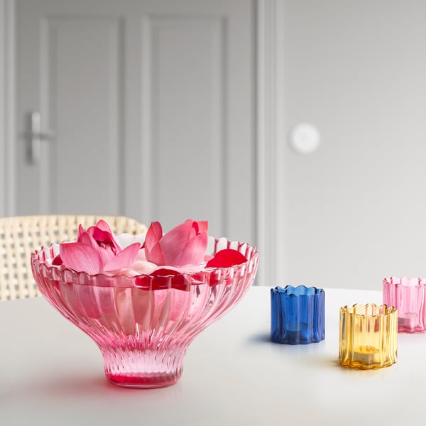 A glass pink AROMATISK serving bowl sitting on a off-white table filled with rose petals