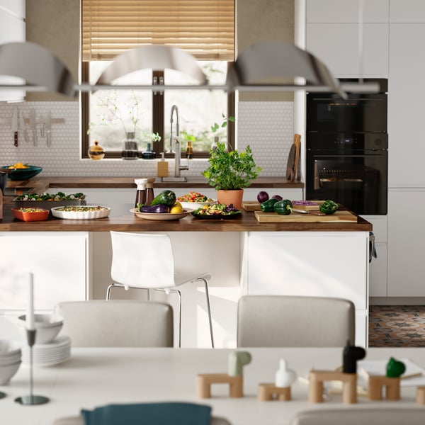 Uma cozinha luminosa com frentes VOXTORP brancas mate e uma península de cozinha, com uma mesa de jantar branca em primeiro plano.