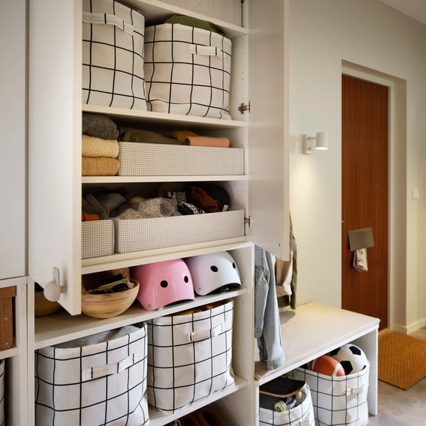 A hallway shelving unit with SLÄTTERVALL storage baskets placed on the shelves alongside helmets and folded textiles.