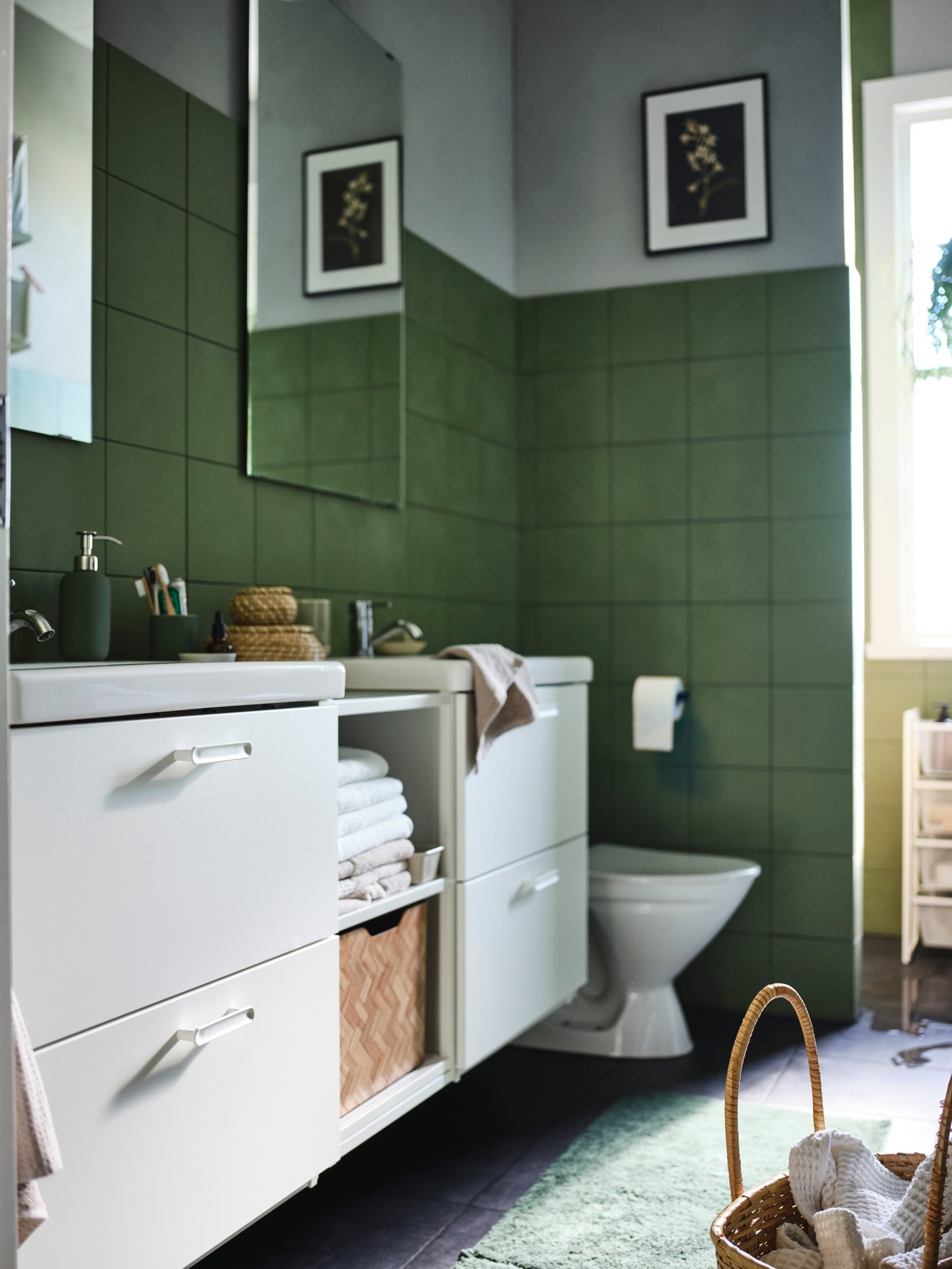 A bathroom with green tiled walls, ENHET bathroom cabinets, mirrors above white sinks, drawers and shelves holding towels.