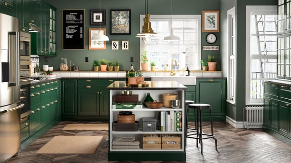 A kitchen in dark green with a kitchen island, black bar stools, a brass-coloured mixer tap and two jute rugs.