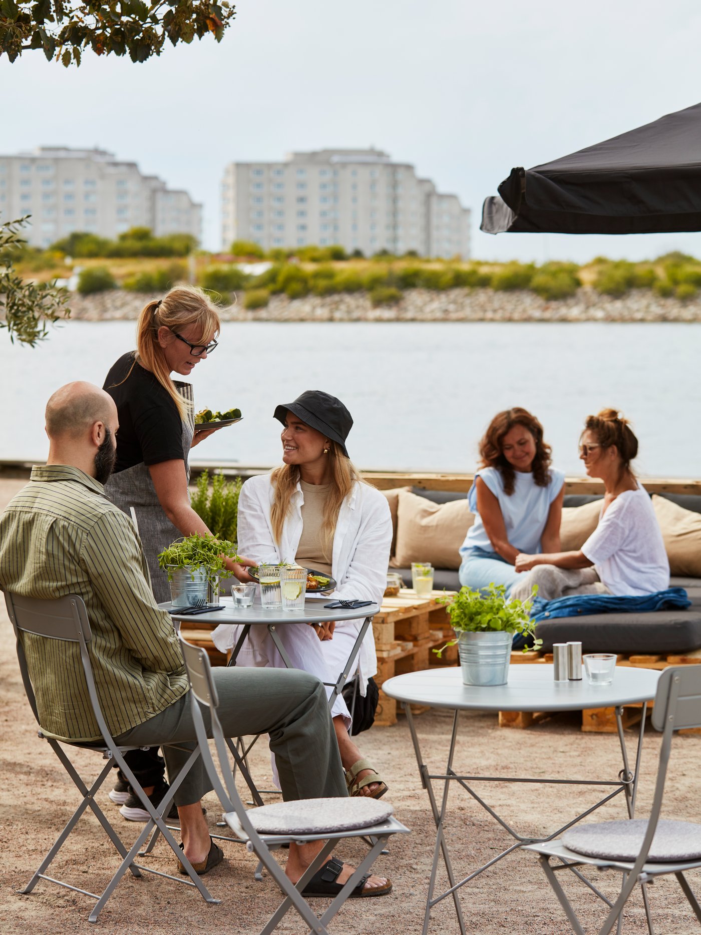 A server attends to customers sitting outside an outdoor café by the water with grey SUNDSÖ outdoor chairs and tables.
