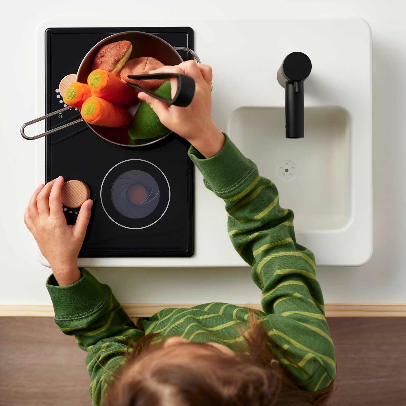 Child playing with a toy stovetop, stirring colorful pretend vegetables in a pot next to a small sink.