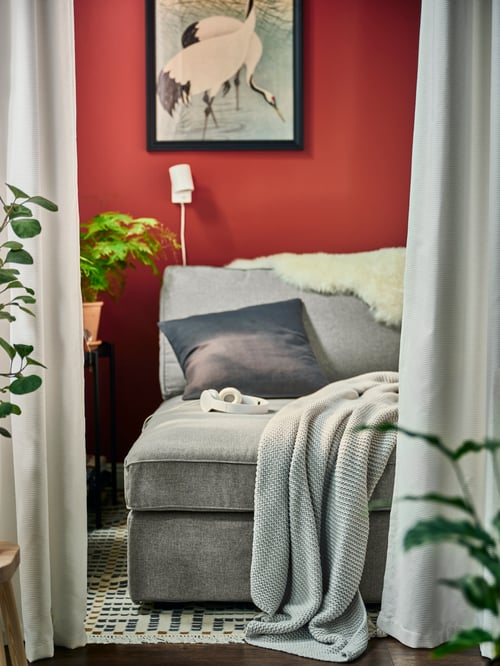 Modernes Wohnzimmer mit roten Wänden und einer KIVIK Récamiere hinter GUNNLAUG geräuschdämmenden Gardinen, u. a. mit einem INGABRITTA Plaid.
