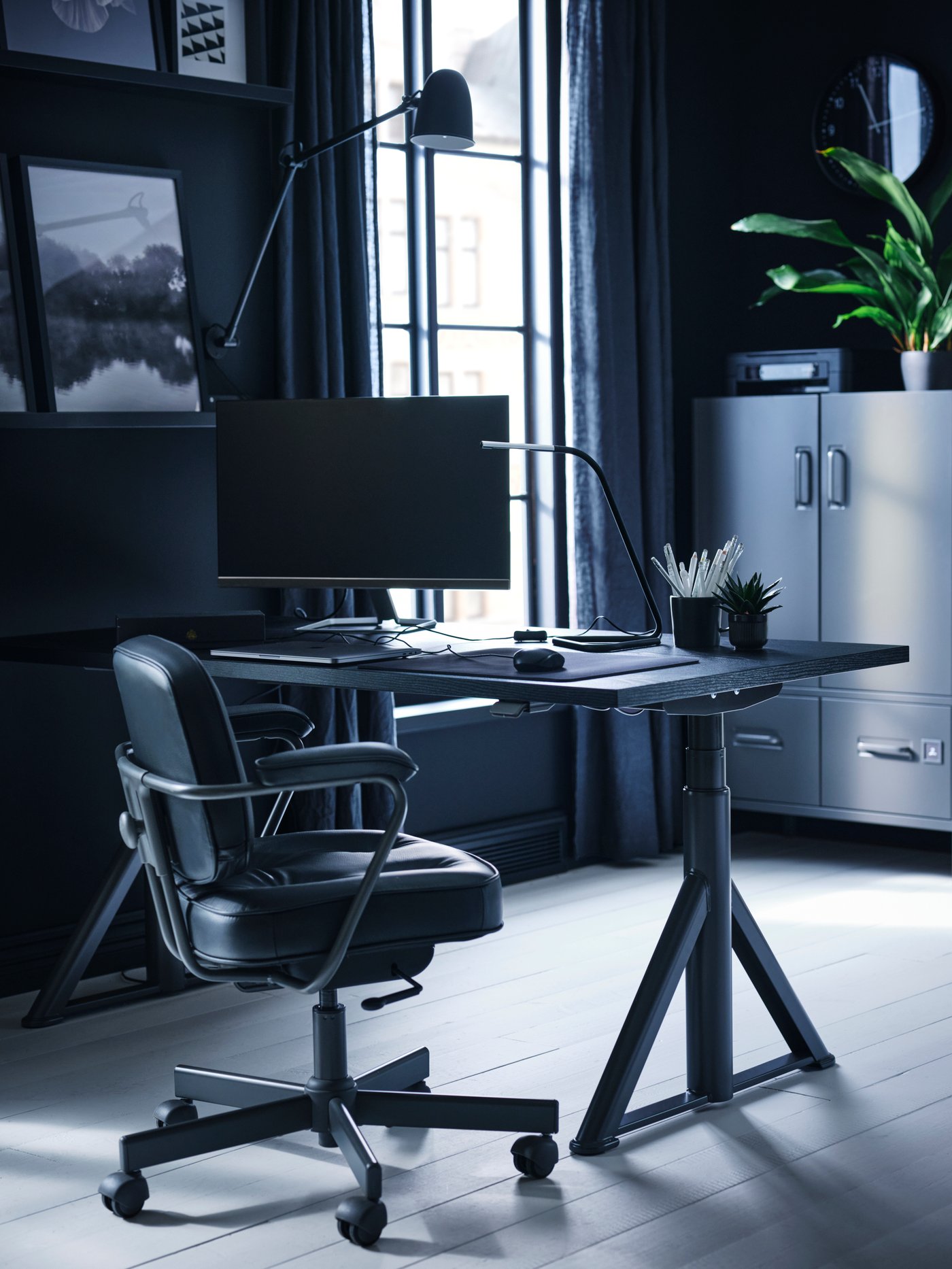 A black/dark grey IDÅSEN sit/stand desk in a home workspace near a window, with pictures in black frames on one wall.