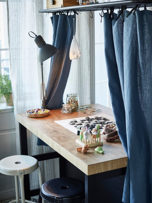 Workspace with kitchen island with rack, bar stools, work lamp, toys, curtains, white curtains, windows, potted houseplants.