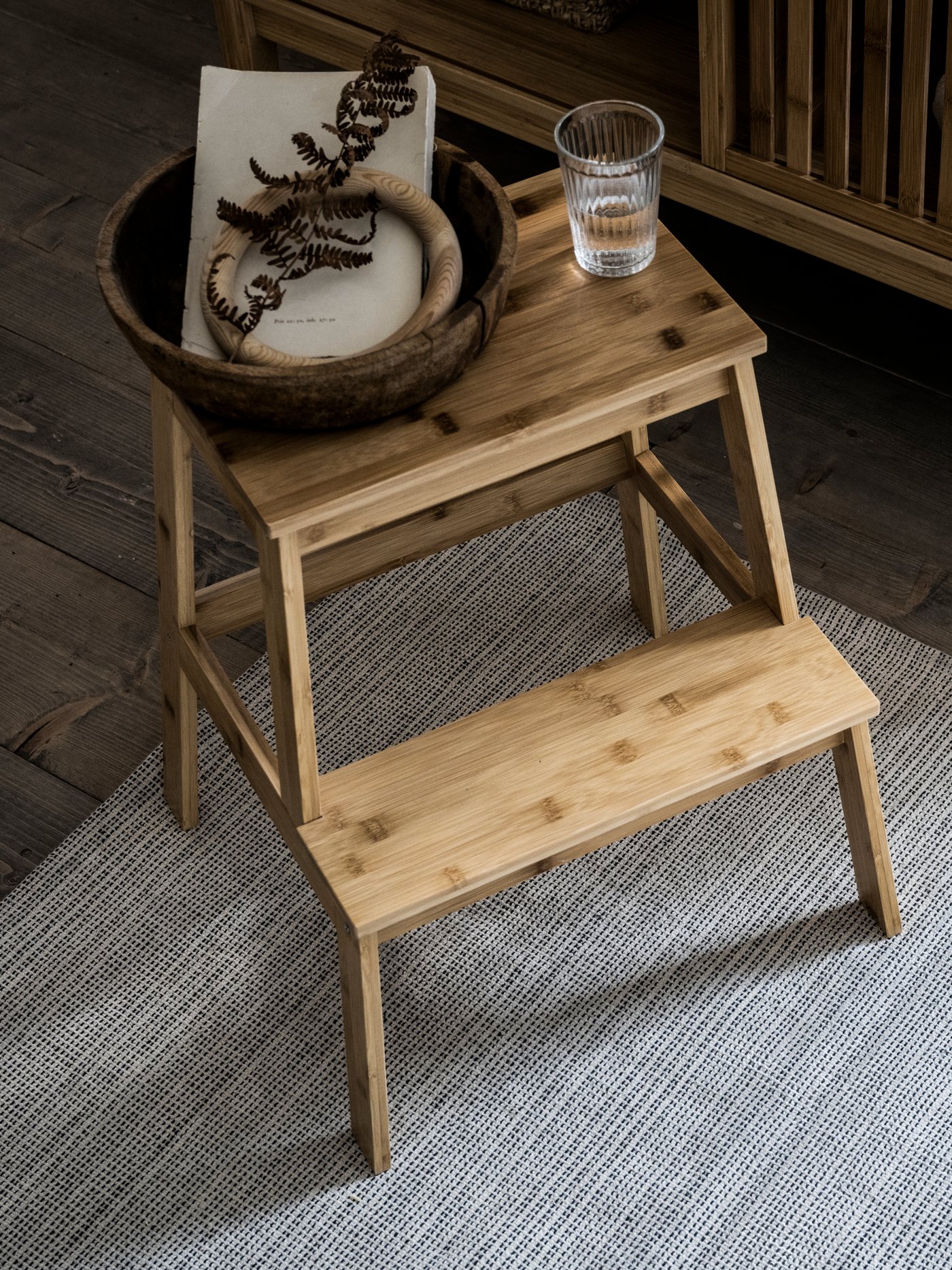 سلم متدرج TENHULT عليه كوب من الماء ووعاء خشبي يحتوي على كتاب وحلقة خشبية وبعض أوراق الشجر الجافة.