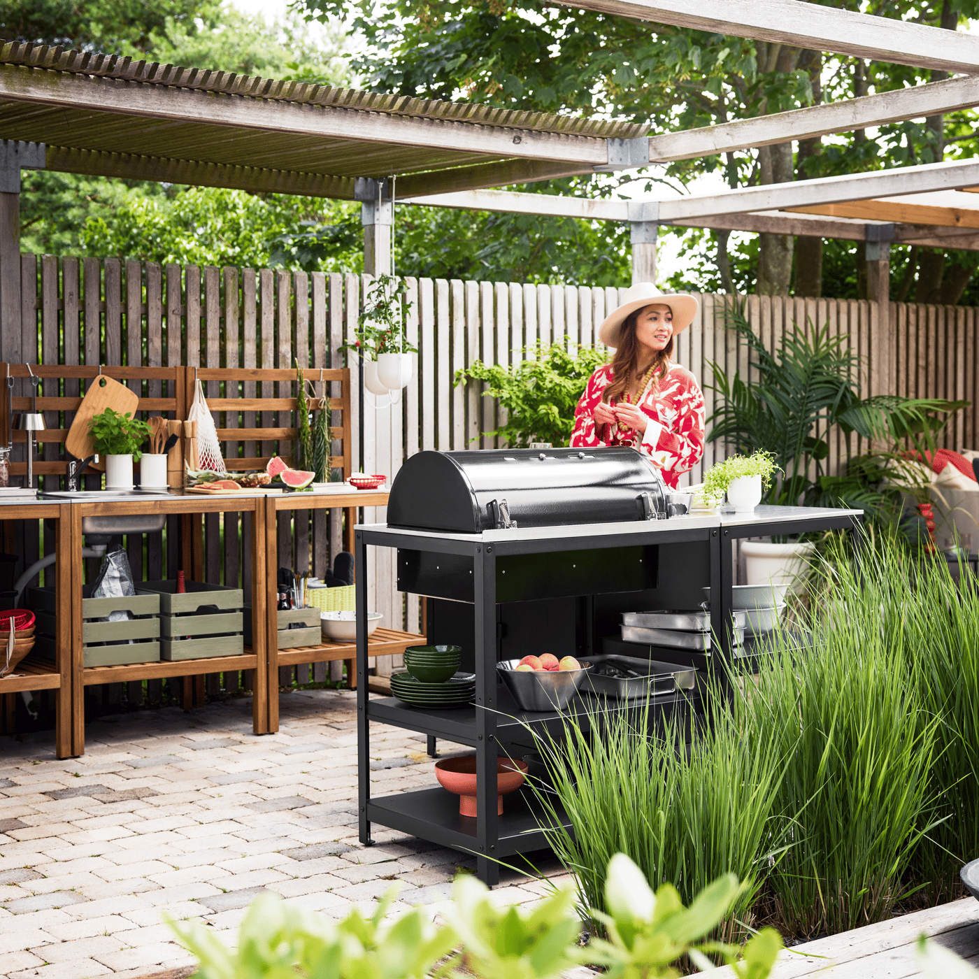 Woman at outdoor grill on patio with pergola and green plants.