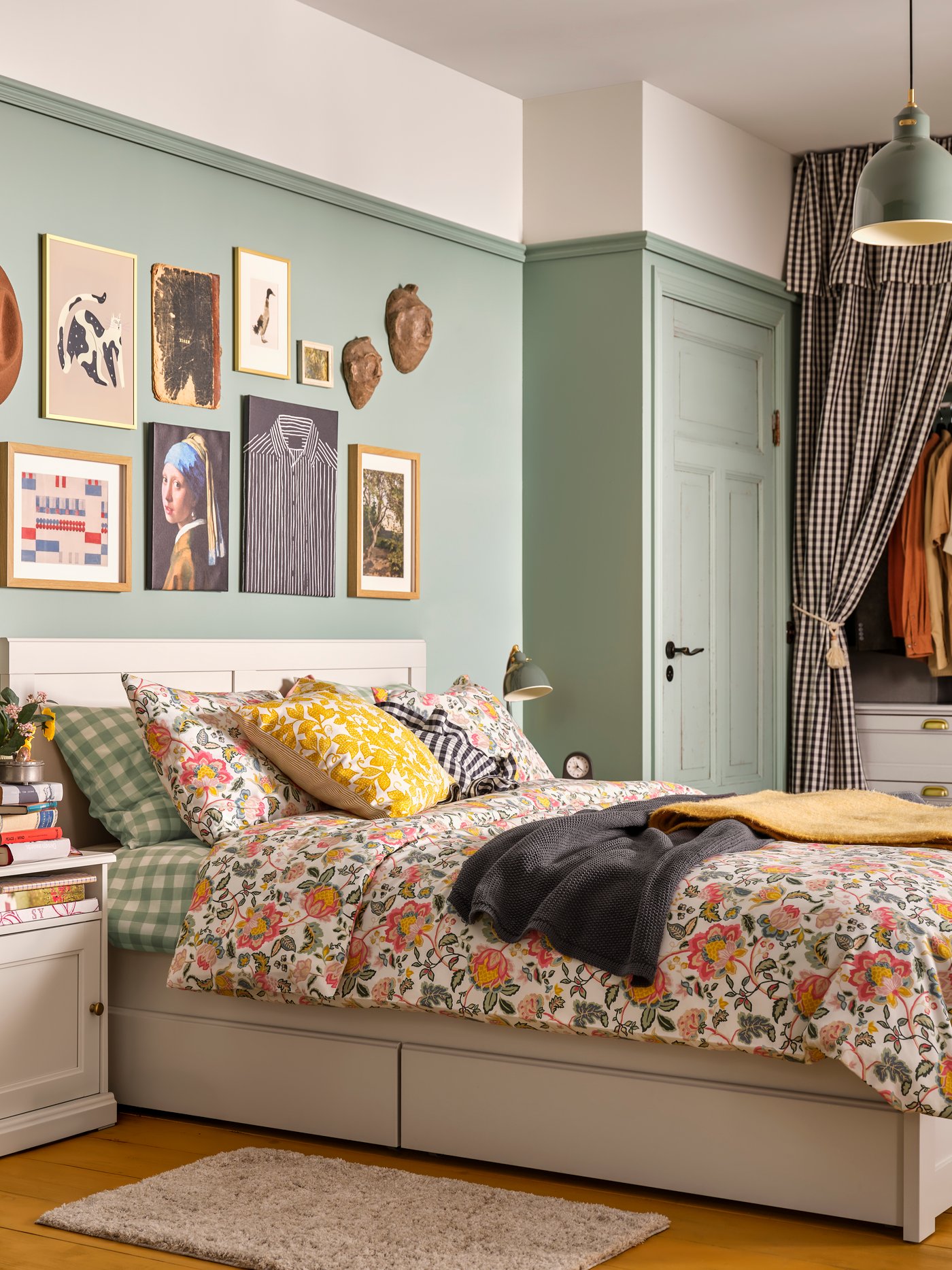 Camera da letto accogliente con letto FÅGELFJÄLLET bianco sporco con cassetti contenitore, biancheria floreale colorata e parete verde decorata con quadri.