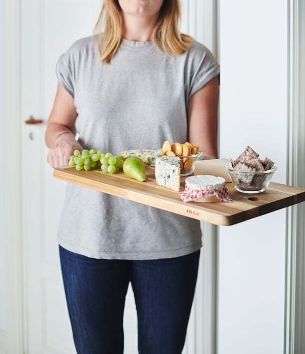 Eine Person in Jeans und T-Shirt, die ein SMÅÄTA Schneidebrett mit Käse und Obst in Händen hält.