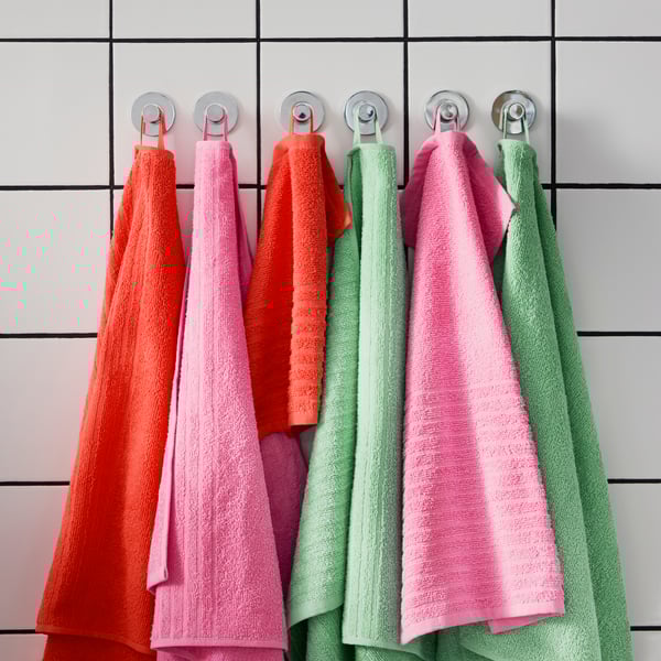 Sis tovalloles de cotó VÅGSJÖN de color verd, rosa i vermell penjades en ganxos a la paret d'un bany amb rajoles blanques.