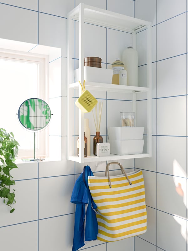 The corner of a modern deco style bathroom, showing a make-up mirror on a windowsill beside open shelving holding simple storage boxes and a striped bag.