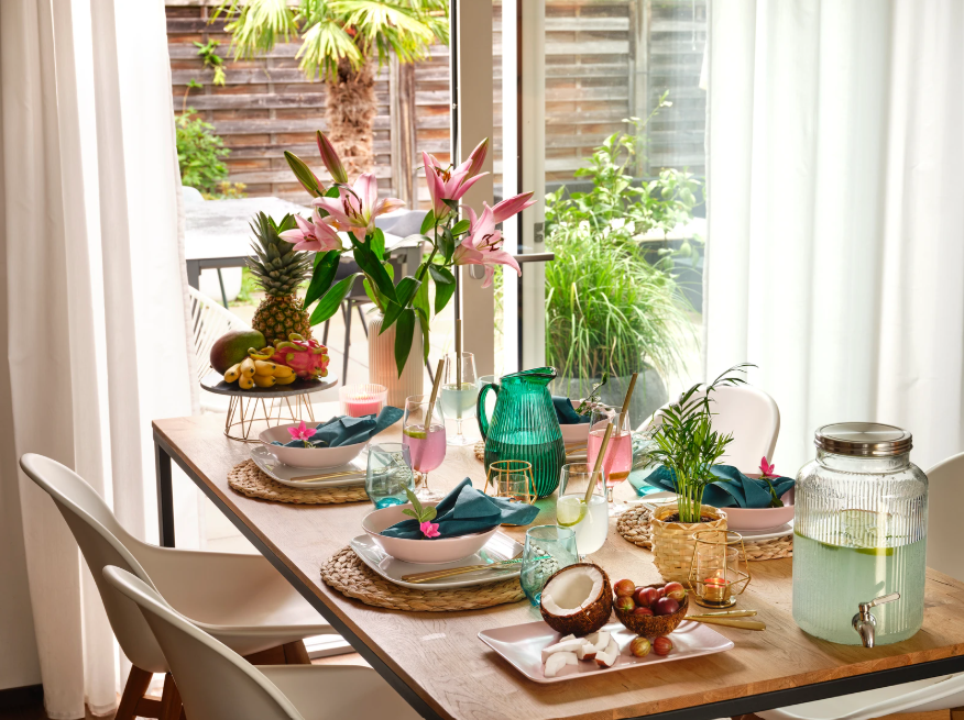 Sommerlich gedeckter Tisch mit farbigem Geschirr, Blumen, Getränkespender und Obst vor grossen Fenstern mit Gartenblick.