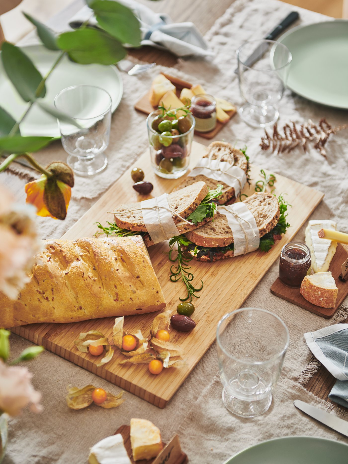 Detalle de una mesa de comedor con comida en una tabla de cortar, platos de gres con esmalte verde mate, copas y cubiertos.