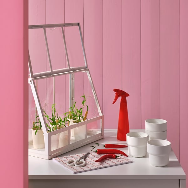 A white table with ÅKERBÄR greenhouse, potted herbs, and a red TOMAT spray bottle against a pink wall.