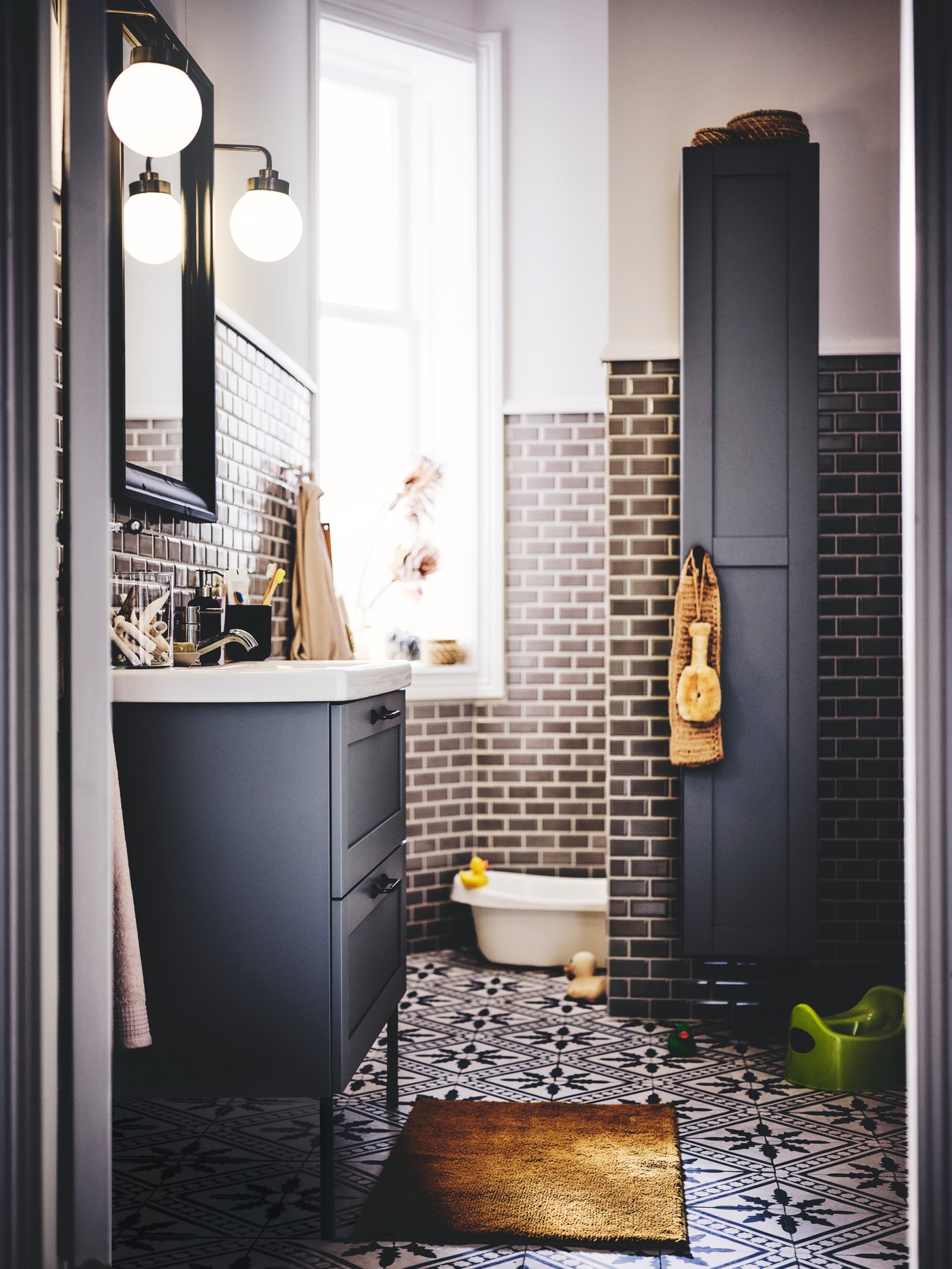 חדר אמבטיה שבו כיור ENHET/TVÄLLEN אפור, מנורות קיר, ארון גבוה, אמבטיית תינוק, שטיח אמבטיה ומגבות.