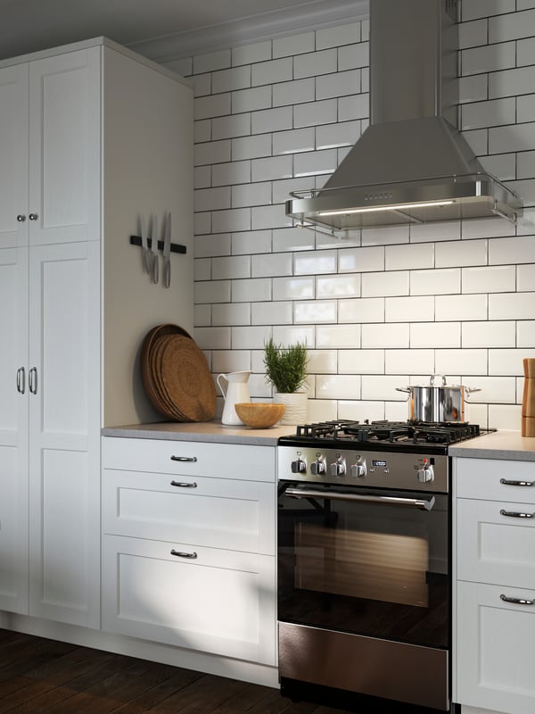 Une cuisine avec des façades ENKÖPING blanches, une hotte murale ALLMÄNBILDAD en acier inoxydable et des objets sur le comptoir.