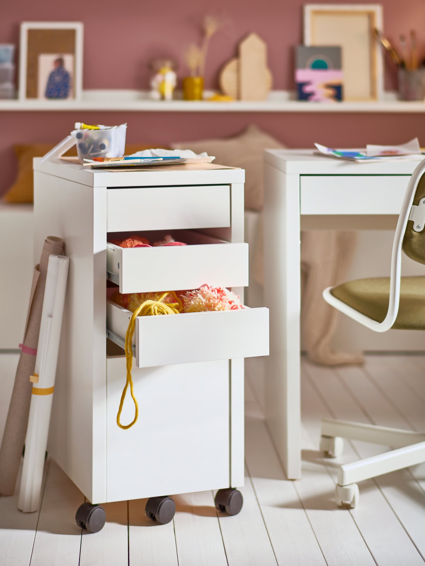 Un módulo de cajones MICKE blanco con espacio para almacenar archivos y dos cajones llenos de hilos, un escritorio infantil y una silla giratoria.