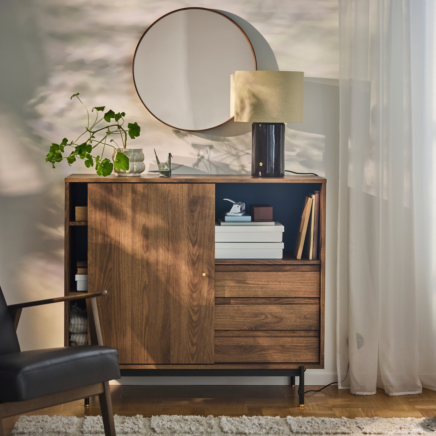 RÅDMANSÖ cabinet in dark wood with books and decor, placed beneath a round mirror in a living room.