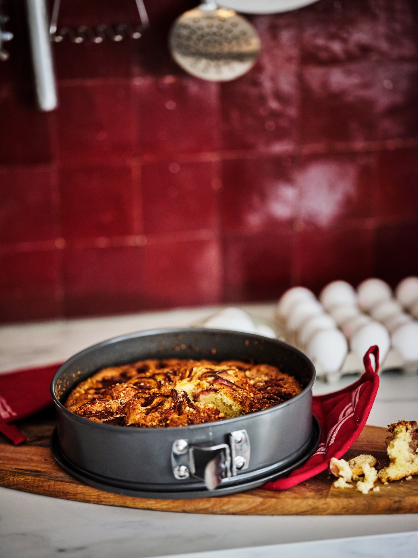 Detailní pohled na kuchyňskou desku s červenými dlaždicemi v pozadí. Na dřevěném prkénku leží upečený koláč ve formě HEMMABAK.
