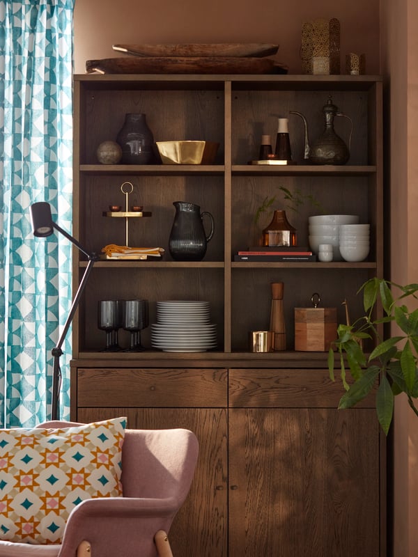 A set of white GOKVÄLLÅ bowls, white GOKVÄLLÅ plates and GOKVÄLLÅ serving tray sitting on a brown wooden cabinet shelves for display in the living room.