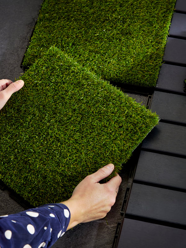 Hands installing a section of floor decking in artificial grass next to dark grey decking.