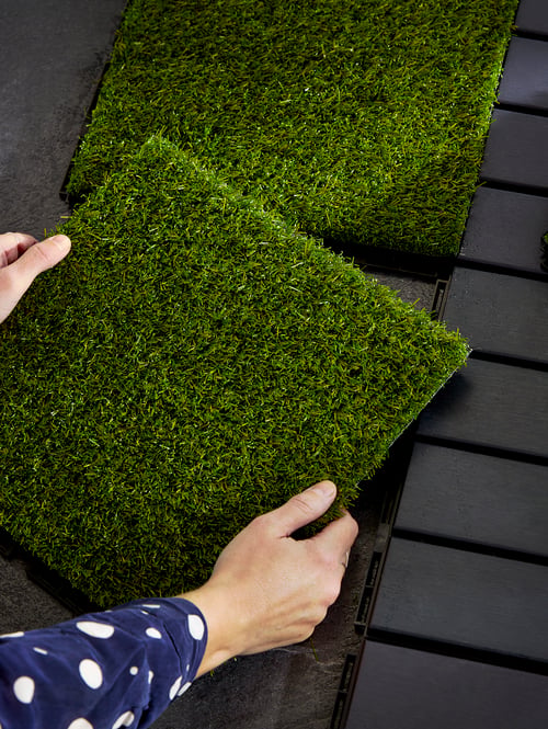 A pair of hands holds a square of RUNNEN artificial grass outdoor floor decking to place it beside other slatted decking.