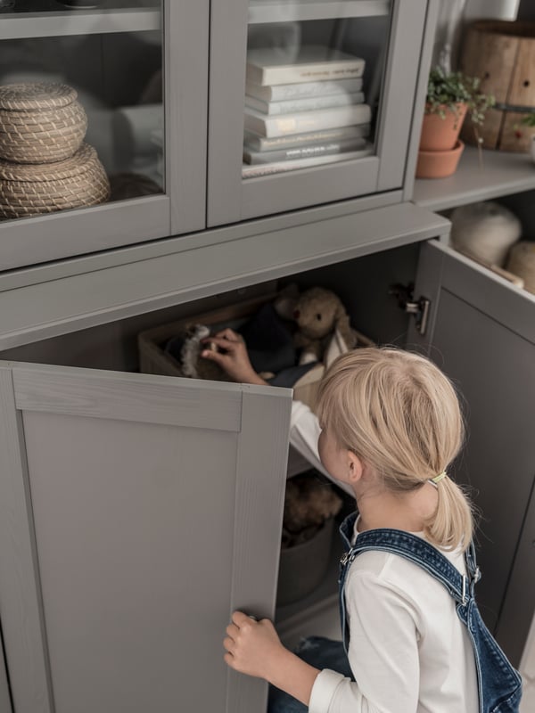 A child with denim overalls opens the bottom doors and reaches into a HAVSTA storage combination with glass doors.