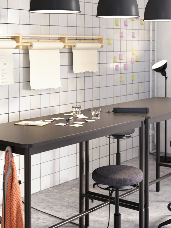 Two black high tables and several black high stools standing together beside a white wall with colourful sticky notes.