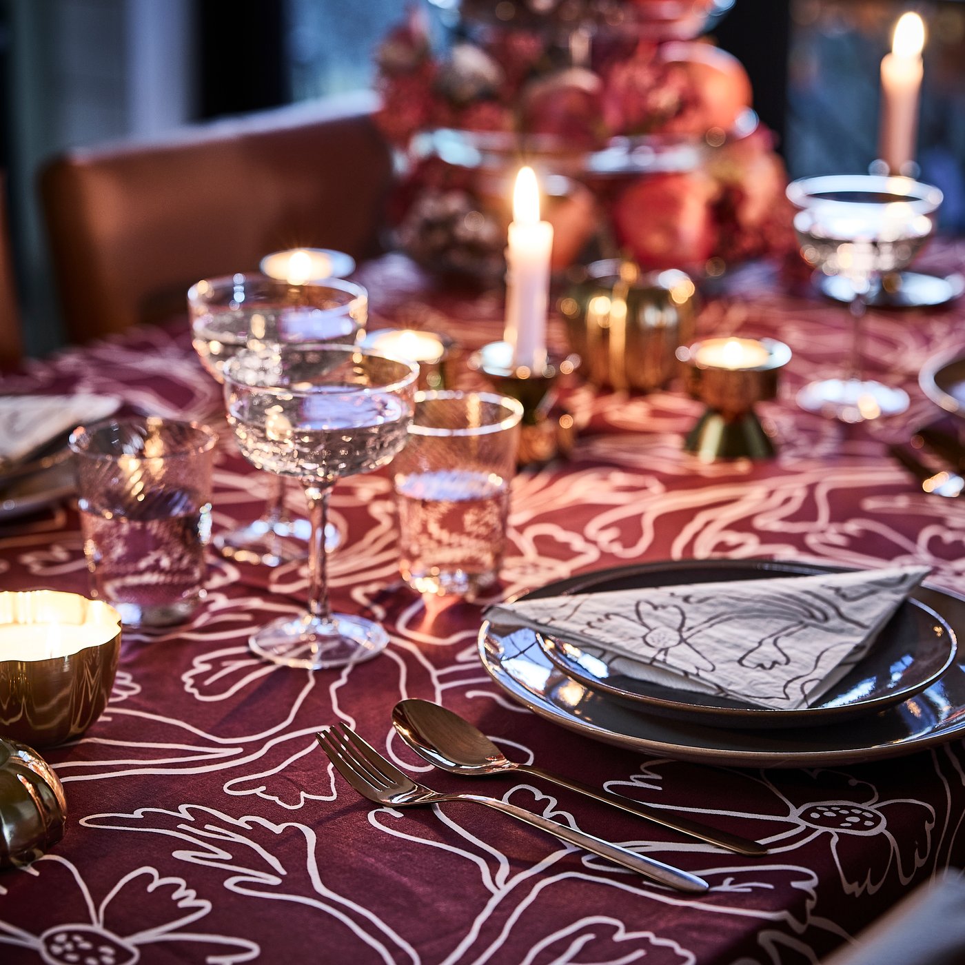 A pink and red table dining table is set with lit candles, a red patterned HÖSTAGILLE tablecloth, paper napkins and coupes.