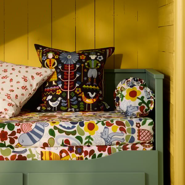 A multicolour TRATTKAKTUS cushion cover placed on a HEMNES day-bed frame with 3 drawers in grey-green against a yellow wall.