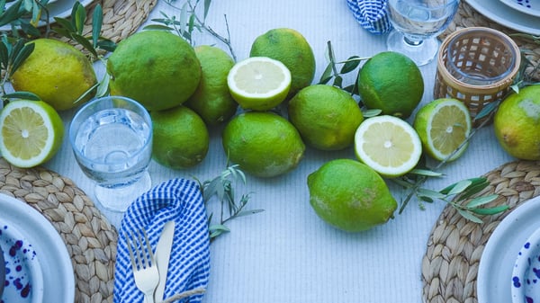 Table dressée avec des assiettes IKEA Silvereda, des couverts NATTKLOT, des verres à pied FRUKTDUVA, une serviette bleue et blanche, des citrons verts et des olives. Idéal pour les repas d'été à la méditerranéenne.