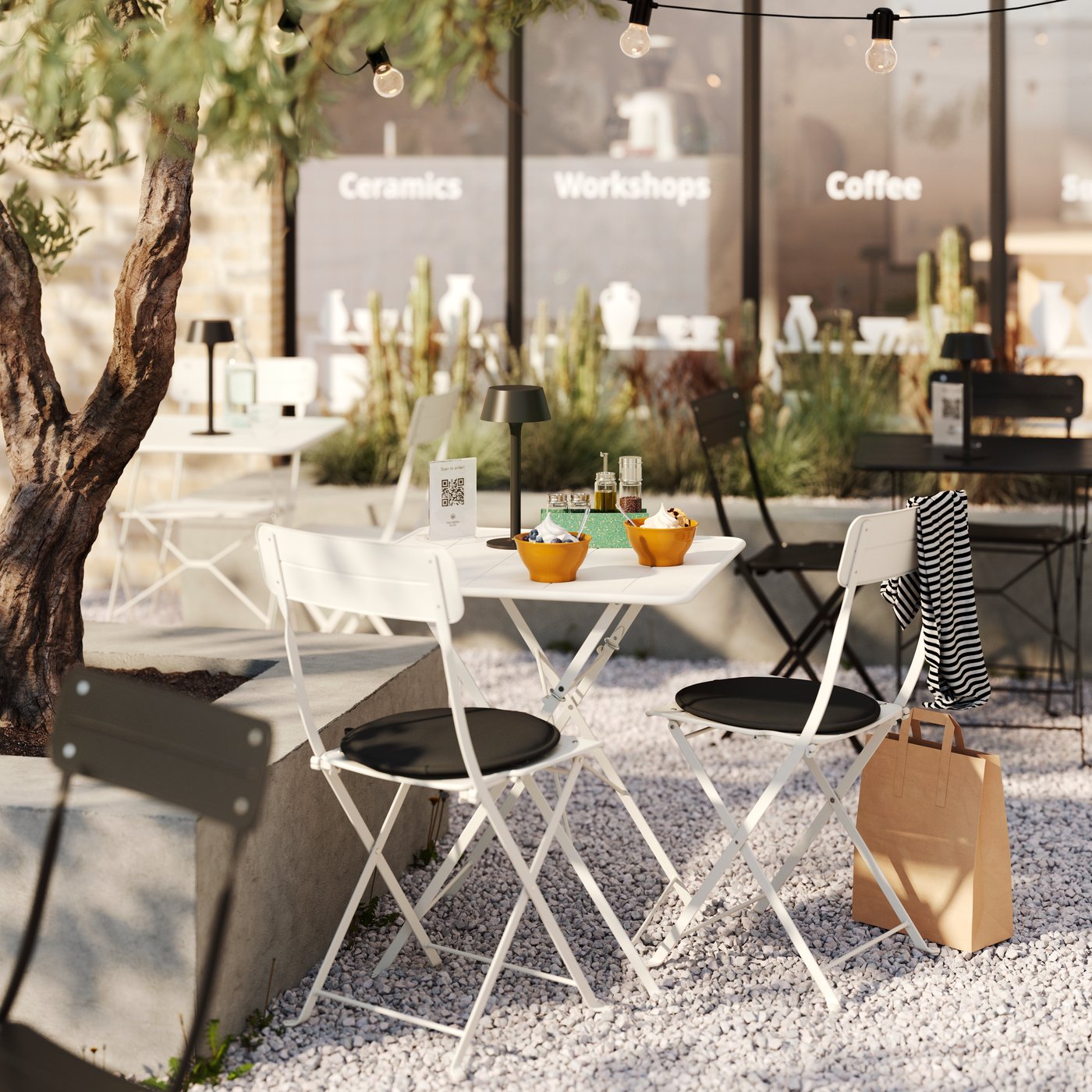 A set of SUNDSÖ folding chairs around a table standing on gravel next to an olive tree in the outdoor service area of a café.