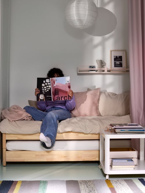 O femeie stă pe două paturi suprapuse UTÅKER din pin, citind o revistă, cu o măsuță auxiliară albă, un covor multicolor și o lustră alături.