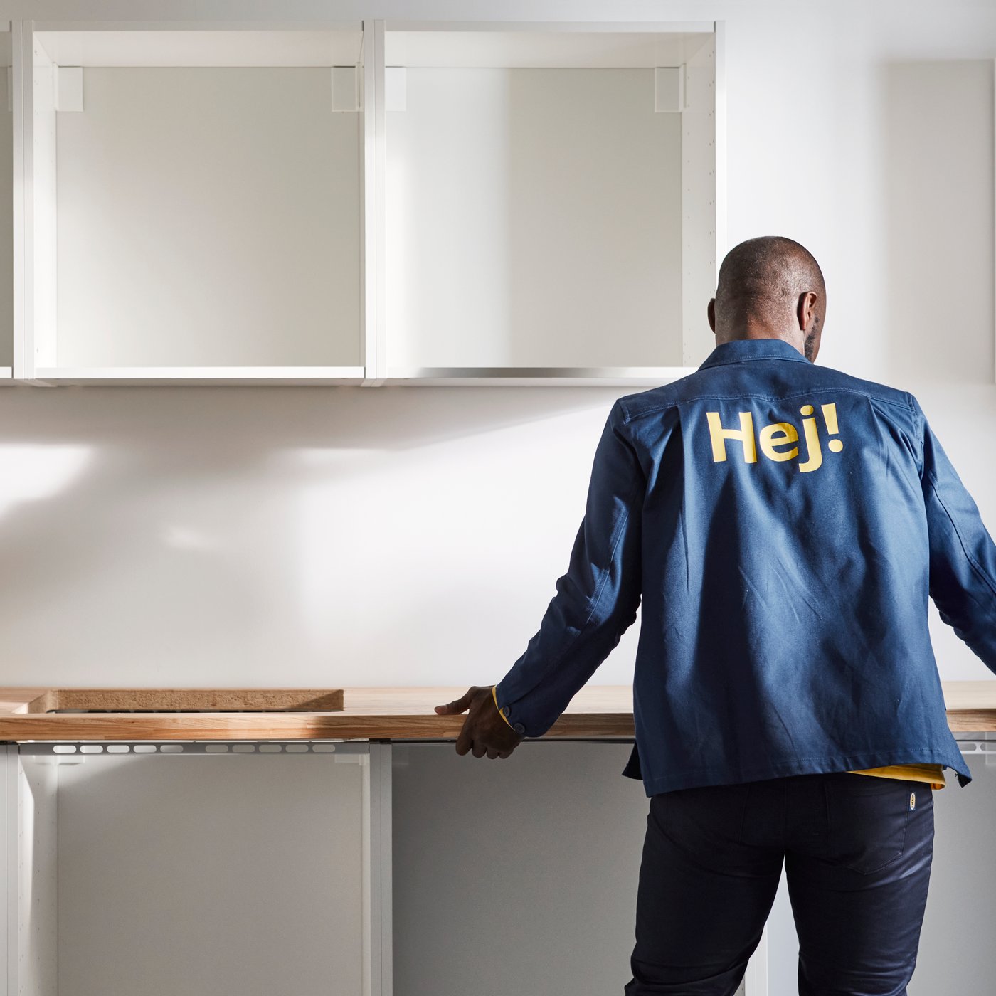 V delno dokončani kuhinji stoji zaposleni podjetja IKEA, ki je obrnjen stran in drži kuhinjsko delovno ploščo.