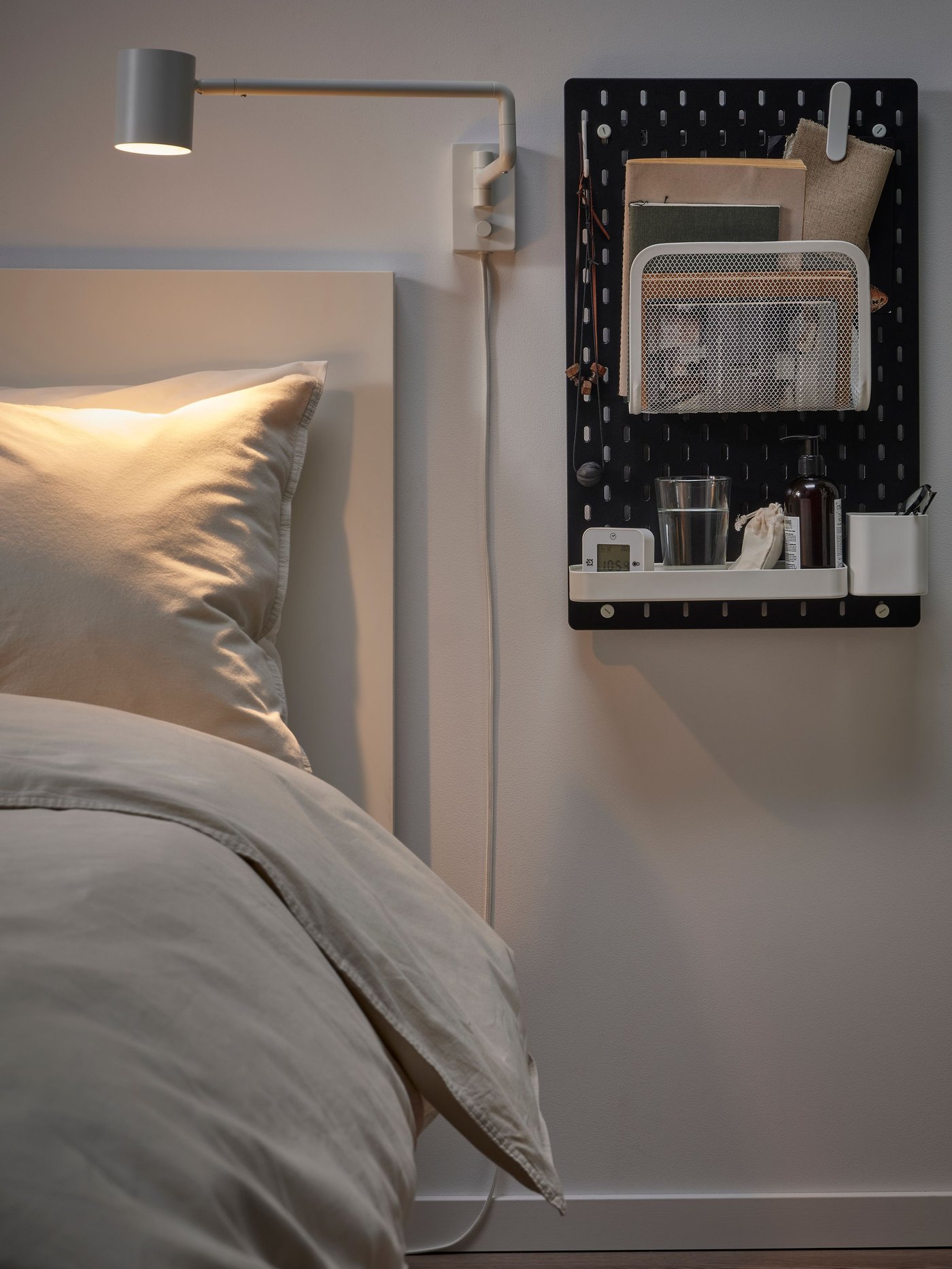 Una habitación con una cama y al costado derecho un tablero SKÅDIS negro en la pared cumpliendo las funciones de velador. 